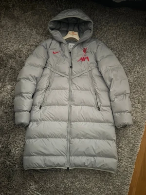 Ljusgrå lång dunjacka Nike Liverpool - Ljusgrå lång dunjacka från Nike med Liverpool FC-logga och röda detaljer. Jackan har huva, två stora dragkedjeförsedda fickor och quiltad design. Perfekt för kalla vinterdagar och visar tydligt ditt stöd för Liverpool.