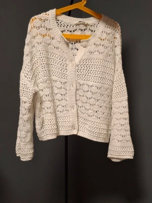 Vit virkad kofta med knappar - Säljer en söt vit stickad kofta med virkat mönster och rund hals. Koftan har knappar framtill och är gjord i ett mjukt, luftigt material. Perfekt att slänga över en topp för en chill look.