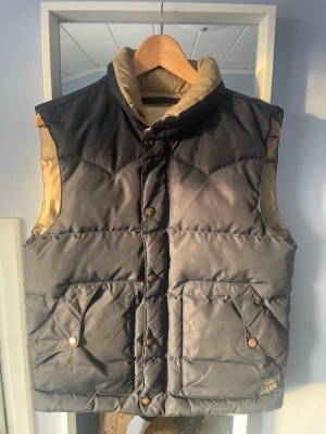 Svart dunväst från Ralph Lauren - Stilren svart dunväst från Denim & Supply Ralph Lauren med beige insida och detaljer. Västen har hög krage, tryckknappar framtill och två stora fickor med lock. Perfekt för lager på lager och ger en klassisk look till din outfit.