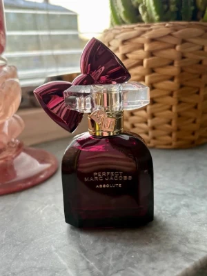 Perfect Marc Jacobs Absolut parfym - En lyxig parfym från Marc Jacobs, Perfect Absolut, i en mörklila glasflaska med en stor dekorativ rosett i plast på korken. Flaskan har gulddetaljer och text i guld. Perfekt för dig som vill ha en unik och snygg parfymflaska på hyllan. 30ml. Ny