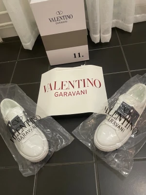 Valentino Garavani vita sneakers - Snygga vita sneakers från Valentino Garavani med svart detalj över snörningen och nitar på sidorna. Skorna har en klassisk låg siluett och är tillverkade i slätt läder. Perfekta för dig som vill ha en stilren och exklusiv look.