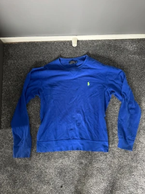 Blå crewneck från Polo Ralph Lauren - Blå crewneck från Polo Ralph Lauren med klassisk rund halsringning och den ikoniska gröna logon broderad på bröstet. Tröjan har långa ärmar och ribbade muddar vid ärmslut och nederkant. Perfekt för en avslappnad och stilren look.