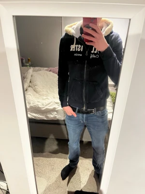 Marinblå hoodie från Hollister - Säljer en marinblå hoodie från Hollister med dragkedja och vit broderad text på bröstet. Hoodien har en mysig, fluffig foder i huvan och vita snören. Perfekt för dig som gillar en avslappnad och stilren look. Storlek M