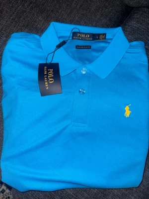 Blå piké från Polo Ralph Lauren - Snygg blå pikétröja från Polo Ralph Lauren med klassisk krage och två knappar framtill. Ikoniska gula loggan broderad på bröstet. Perfekt för dig som vill ha en stilren och tidlös look.
