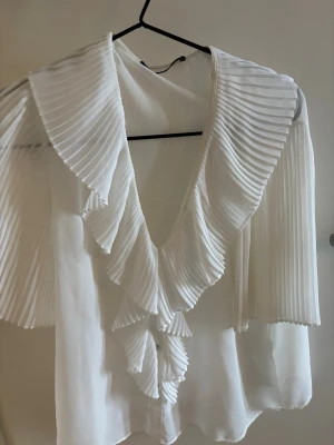 Vit plisserad blus från Zara - Trendig vit blus från Zara med stora plisserade volanger längs kragen och ärmarna. Blusen har en djup V-ringning och är tillverkad i ett lätt, flowy material som ger en elegant look. Perfekt för dig som gillar statement-detaljer och stilrena plagg.