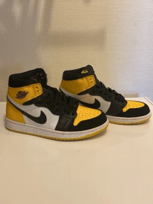 Nike Air Jordan 1 Mid svart/gul - Säljer ett par Nike Air Jordan 1 Mid sneakers i svart, gul och vitt. Klassisk high-top modell med svart Swoosh, gula detaljer på tå, häl och skaft samt vita sidopaneler. Skorna har svarta skosnören och perforerad tåbox för extra ventilation. Perfekta för dig som gillar streetwear.
