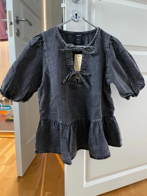 Svart denimblus med puffärm från Lindex - Cool svart denimblus från Lindex med puffiga korta ärmar och volang nedtill. Blusen har två dekorativa knytband framtill och en avslappnad passform. Materialet är tvättad denim som ger en trendig look.
