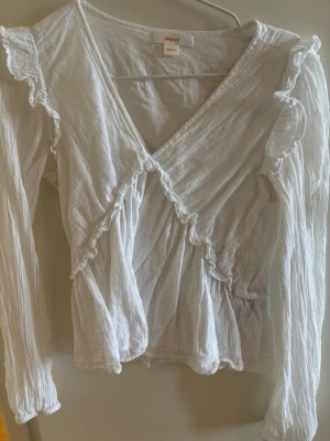 Vit omlottblus med volanger Gina Tricot - Söt vit omlottblus från Gina Tricot med långa ärmar och volangdetaljer längs axlar och omlottringning. Tillverkad i ett lätt, krinklat tyg som ger en avslappnad men ändå chic look. Perfekt att styla med jeans eller kjol för en trendig outfit.