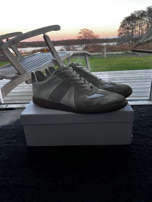 Maison Margiela Skor - Snygga sneakers i grått och vitt med ovandel i mocka och släta läderpaneler. Klassisk låg modell med mörk sula och vita skosnören. Perfekta för dig som gillar stilrena och tidlösa sneakers. Skick 9/10, inga defekter. Storlek 44. Allt på bilderna ingår. Äktahetsbevis saknas. Pris går att diskutera✅