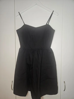 Svart kort klänning med tunna band - En svart kort klänning med tunna axelband och markerad midja. Klänningen har en enkel och stilren design med lätt utställd kjol. Perfekt för dig som gillar minimalistisk stil.