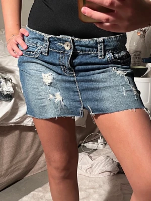 Jeanskjol med låg midja - Säljer en blå jeanskjol med rå kant och slitna detaljer framtill. Kjolen har klassiska fickor, bälteshällor och vit knapp. Perfekt för dig som gillar en avslappnad och trendig stil.
