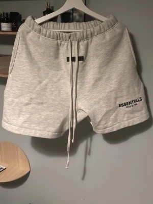 Essentials shorts  - Snygga ljusgrå shorts från Essentials Fear of God med svart logga på vänster ben. De har elastisk midja med långa snören och är tillverkade i mjuk bomull. Perfekta för en avslappnad och trendig look.