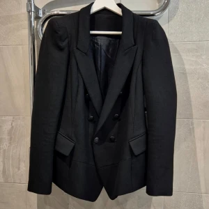 Svart dubbelknäppt kavaj - Stilren svart kavaj med dubbelknäppning och markerade axlar. Kavajen har två fickor med lock framtill och en figurnära passform. Lapp i nacke bortklippt. Klassisk krage och slits bak för extra rörelsefrihet. Perfekt för en dressad look.