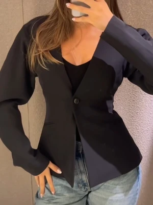 Svart kavaj med v-ringning från Gina Tricot - Stilren svart kavaj från Gina Tricot med djup v-ringning och en knapp framtill. Kavajen har långärmade ärmar och en figurnära passform som ger en snygg siluett. Perfekt att matcha med jeans eller kjol för en trendig look. Storlek L. 