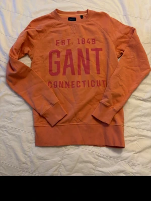Orange sweatshirt från GANT - Säljer en orange sweatshirt från GANT med tryck i rosa text 'EST. 1949 GANT CONNECTICUT' på bröstet. Tröjan har rund halsringning, ribbade muddar vid ärmar och nederkant samt en avslappnad passform. Perfekt för en chill och sportig look.