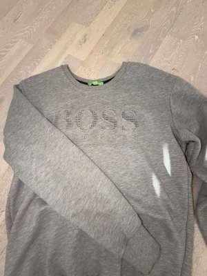 Grå sweatshirt Hugo Boss L/M - Snygg grå sweatshirt från Hugo Boss med rund halsringning och ribbade muddar. Framsidan har ett stort BOSS-tryck med diskret mönster i bokstäverna. Perfekt till jeans eller joggers för en clean och stilren look.