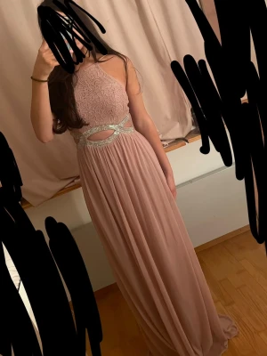 Lång rosa spetsklänning med glitterdetaljer - Elegant långklänning i ljusrosa med spetsliv och glittriga band i midjan. Klänningen har hög hals, är ärmlös och har en öppen detalj vid midjan. Kjolen är i ett mjukt, flytande tyg som ger en romantisk känsla. Perfekt för bal eller festliga tillfällen. Köpt i USA