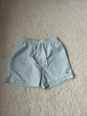 Ljusblåa badbyxor från Polo Ralph Lauren - Snygga ljusblåa badbyxor från Polo Ralph Lauren med elastisk midja och vit snörning. Klassisk logga broderad i mörkblått på vänster ben. Badbyxorna har sidofickor och en bakficka med lock. Perfekta för sommardagar och strandhäng.
