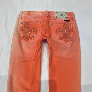 Korallfärgade Miss Me jeans med strass - Snygga orange jeans från Miss Me med raka ben och dekorativa bakfickor prydda med stora fleur-de-lis-mönster i strass och nitar. Jeansen har klassisk femficksmodell och cool tvättad look. Obs inringade fläckar 