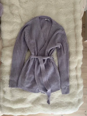 Lila stickad kofta med snöre runt midjan - Lila stickad kofta med snöre runt midjan. Knappt använd. Parfekt för våren eller varför inte till en sen sommarkväll .  