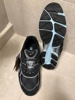 Svarta och grå Asics sneakers - Snygga sneakers från Asics i svart och grått med meshpaneler och detaljer i ljusblått. Skorna har en sportig design med robust sula och klassisk snörning. Perfekta för dig som gillar streetwear och vill ha bekväma skor med cool look.