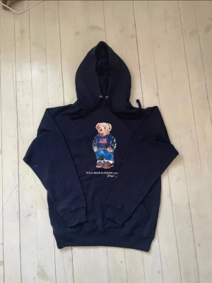 Mörkblå hoodie Polo Bear Ralph Lauren - Säljer en mörkblå hoodie från Ralph Lauren med den ikoniska Polo Bear-trycket framtill. Hoodien har huva med dragsko och en stor magficka. Materialet är mjukt och skönt, perfekt för en avslappnad stil. Klassisk design med amerikansk flagga på björnens tröja.