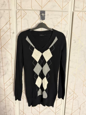 Svart stickad tröja med argylemönster - Snygg svart stickad tröja från Equipe med klassiskt argylemönster i vitt och grått framtill. Tröjan har V-ringning, långa ärmar och ribbade muddar vid ärmslut och nederkant. Ett litet hål under ena armhålan men som man inte direkt märker av