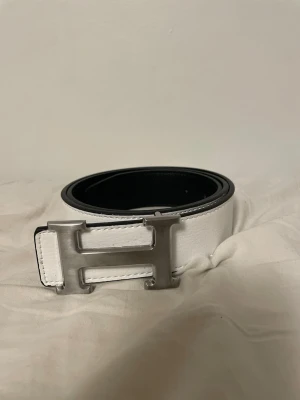 Vitt skärp med H-spänne - Snyggt vitt skärp med ett stort, silverfärgat H-spänne. Skärpet har en stilren design med svart insida och är tillverkat i skinn. Perfekt accessoar för att lyfta vilken outfit som helst. 