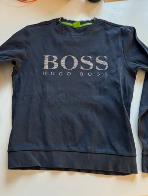 HUGO BOSS mörkblå tröja - Mörkblå sweatshirt från Hugo Boss med stort BOSS-tryck och texten HUGO BOSS på bröstet. Tröjan har rund halsringning, långa ärmar och ribbade muddar vid ärmslut och nederkant. Perfekt för en avslappnad och stilren look.