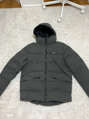 Mörkgrå pufferjacka från Sail Racing - Säljer en mörkgrå pufferjacka från Sail Racing med huva och dragkedja framtill. Jackan har två snedställda fickor med dragkedja och diskret logga på bröstet. Perfekt för kalla dagar och riktigt snygg streetstil. Pris kan diskuteras.