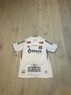 Vit Santos Neymar Jr. matchtröja - Säljer en vit Santos FC matchtröja från Umbro med Neymar Jr. och nummer 10 på ryggen. Tröjan har flera sponsorer tryckta fram och bak, korta ärmar och rund hals. Perfekt för dig som älskar brasiliansk fotboll och vill ha en ikonisk supportertröja.