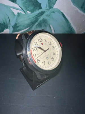 Tommy Hilfiger beige klocka - Stilren klocka från Tommy Hilfiger med beige urtavla, svarta siffror och detaljer i rött och blått. Boetten är i silverfärgad metall och armbandet är i brunt skinn. Perfekt accessoar för dig som gillar klassisk design.