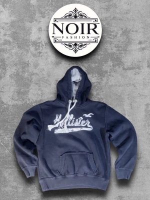 Mörkblå Hollister hoodie - Snygg mörkblå hoodie från Hollister med vit logotyp och fågeltryck på bröstet. Klassisk huva med vita snören och stor magficka framtill. Perfekt för dig som gillar en avslappnad och cool stil.