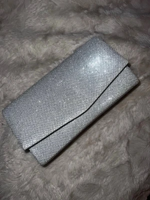 Silver Glitter Clutch Bag  - Elegant silver clutch bag with a beautiful glittery texture that catches the light perfectly. This bag is ideal for special occasions such as parties, weddings, or prom, adding a touch of sparkle to any outfit. Compact yet spacious enough for your essentials like phone, keys, and makeup. Easy to carry and style with both formal and festive looks.