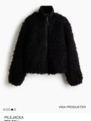 Svart teddyjacka med dragkedja - Mysig svart teddyjacka med fluffig pilestruktur och hög krage. Jackan har en rak passform och stängs med en svart dragkedja framtill. Perfekt för att hålla värmen under kyliga dagar och ger en cool look.