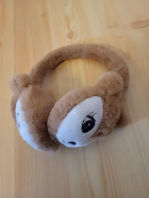 Bruna fluffiga öronmuffar med djurmotiv - Säljer ett par söta bruna öronmuffar i mjuk plysch med djuransikten och små öron på sidorna. Perfekt accessoar för att hålla öronen varma och ge en gullig touch till din outfit. Inte helt säker men tror att jag köpte dem på lindex.
