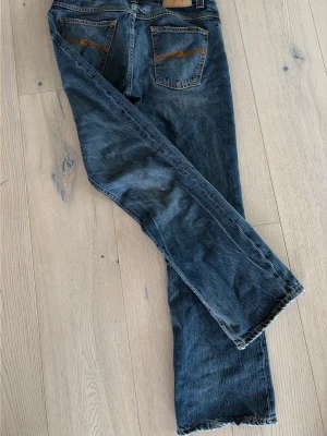 BOOTCUT OLA NUDIE JEANS - Säljer ett par klassiska blå jeans från Nudie Jeans i den sällsynta modellen bootcut Ola.. Jeansen har två bakfickor med den ikoniska Nudie-bågen i orange tråd och en patch bak i midjan. Perfekta för en avslappnad och trendig look.
