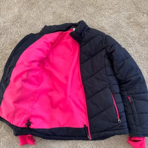 Mörkblå jacka med rosa detaljer - Snygg mörkblå jacka från Everest med rosa dragkedjor och foder. Jackan har hög krage, två fickor framtill, liftkortficka på arm och tumhål i ärmsluten. Perfekt för kalla vinterdagar.