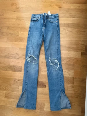 Blå slitna jeans med slits - Säljer ett par blå jeans med slitningar över knäna och höga slitsar vid bensluten. Klassisk femficksmodell med raka ben och normal midja. Perfekta för en avslappnad och trendig look.