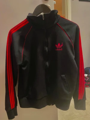 Svart Adidas Originals kofta med röda ränder - Säljer en svart hoodie från Adidas Originals med dragkedja och röda detaljer. Tröjan har de klassiska tre ränderna längs ärmarna och Adidas logga både fram och bak i rött. Tillverkad i mjukt material och har ribbade muddar vid ärmslut och nedkant 