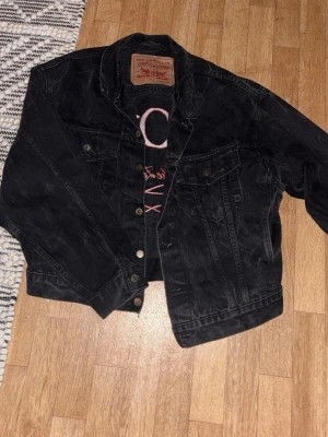 Svart Levi's 501 jeansjacka - Säljer en svart jeansjacka från Levi's med stor röd brodyr på ryggen där det står '501 Live the Legend'. Klassisk modell med knappstängning, två bröstfickor och cool vintagekänsla. Perfekt för dig som vill sticka ut med en unik jacka.