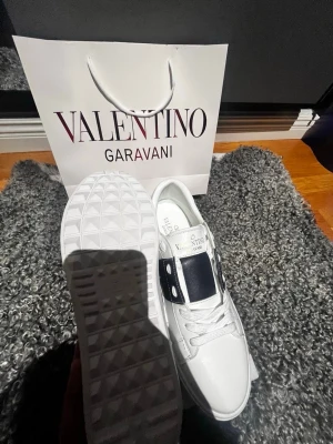 Valentino Garavani vita sneakers - Snygga vita sneakers från Valentino Garavani med svart band och silverdetaljer på sidan. Skorna har en klassisk låg siluett, vita snören och en robust sula med rutmönster undertill. Tillverkade i läder för en lyxig känsla och stilren look. Helt nya, pris kan sänkas 