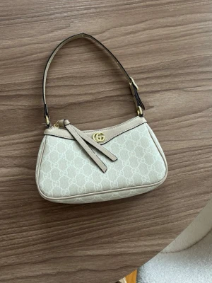 Monogram väska  - Snygg beige axelremsväska från Gucci med klassiskt GG-mönster och guldfärgad logga framtill. Väskan har en stilren form, dragkedja upptill och detaljer i läder. Perfekt för dig som gillar exklusiva accessoarer och vill ha något trendigt.
