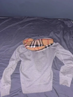 Grå hoodie med Burberry-mönstrad huva - Snygg grå zip-hoodie från Burberry med klassiskt rutmönster i beige, svart och rött på insidan av huvan. Hoodien har dragkedja framtill, fickor och ribbade muddar. Perfekt för en stilren och trendig look.