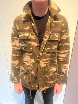 Camouflage vindjacka med snörning - Snygg camouflage vindjacka med flera fickor och snörning i midjan. Jackan har hög krage, knapp- och dragkedjestängning samt patch med stjärna på ärmen. Perfekt för dig som vill ha en streetwear-look. Passar M perfekt