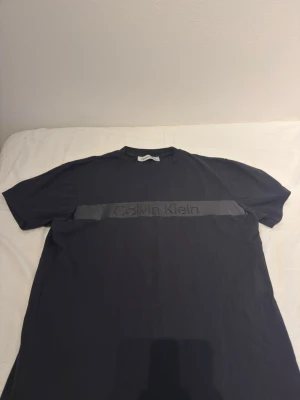 Svart Calvin Klein t-shirt med logga - Snygg svart t-shirt från Calvin Klein med en bred, blank rand över bröstet och stor logga i samma ton. Klassisk passform och rund halsringning. Perfekt för dig som gillar stilrena och moderna plagg.s och m