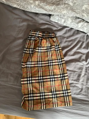 Beige rutiga shorts från Burberry - Säljer ett par klassiska beige shorts från Burberry med det ikoniska rutiga mönstret i svart, vitt och rött. Shortsen har elastisk midja för skön passform och är tillverkade i ett lätt och svalt bomullsmaterial. Perfekta för en trendig och avslappnad stil.