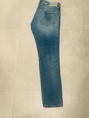 Replay jeans - Säljer ett par blå slim fit jeans från Replay med klassisk femficksdesign och snygga slitningar. Jeansen har normal midja, dragkedjegylf och är tillverkade i stretchigt denim för skön passform. Perfekta till en avslappnad look.
