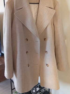 Kappahl jacka 🫶🏽 - Elegant mellanlång beige dubbelknäppt jacka med breda slag i mjuk ull.Klassisk design som passar perfekt till höst 🍂 vinter ❄️ eller vår 🌼Aldrig använd 🎀