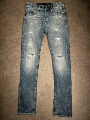 Blå slitna jeans från Jack & Jones 29/32 - Säljer ett par blå jeans från Jack & Jones med slitningar och distressed detaljer framtill och baktill. Jeansen har normal passform, klassisk femficksdesign och snygga kontrastsömmar på bakfickorna. Materialet är jeans i bomull.